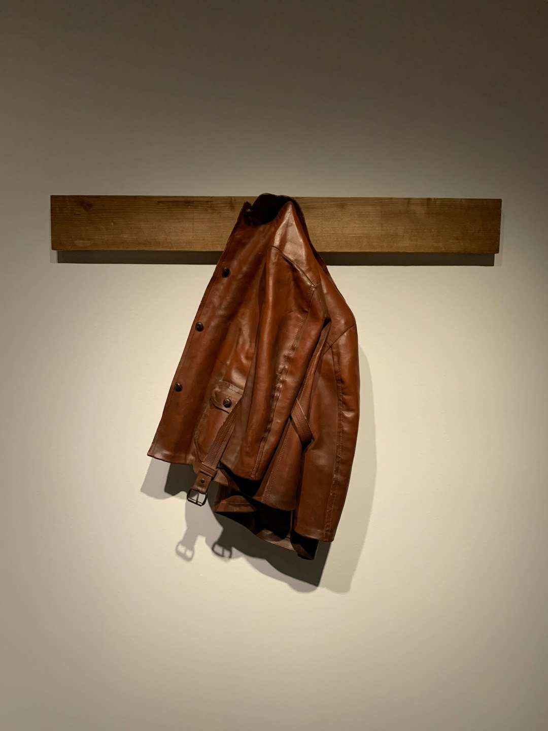 Brown leather jacket with a belt hanging against a neutral wall